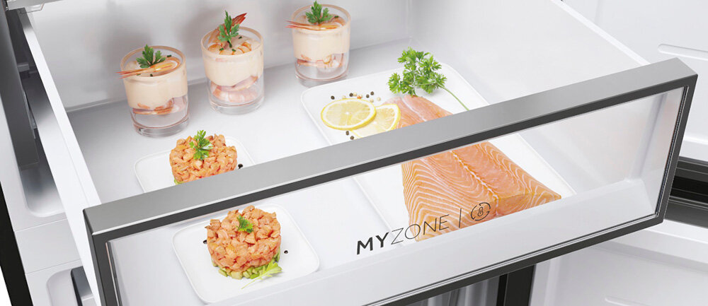 Wnętrze szuflady lodówki z przezroczystym frontem, na którym widnieje napis \'MYZONE\'. W środku znajdują się talerze z łososiem, plasterkami cytryny, ziołami oraz deserami w szklankach.