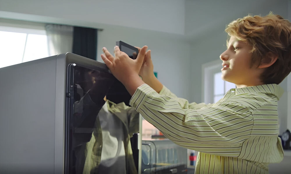 Dziecko w jasnej koszuli obsługuje urządzenie z przezroczystą osłoną, prawdopodobnie drukarkę 3D, w jasnym pomieszczeniu z oknem w tle.