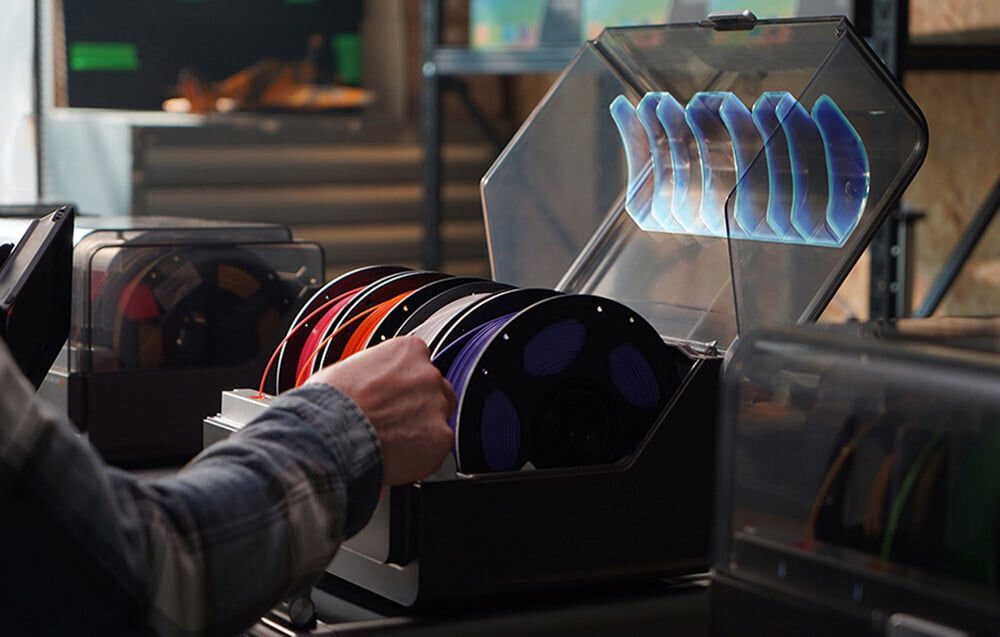 Osoba sięga po szpulę filamentu z otwartego pojemnika, w którym znajduje się kilka kolorowych szpul. W tle widoczne są inne pojemniki z filamentami na półkach.
