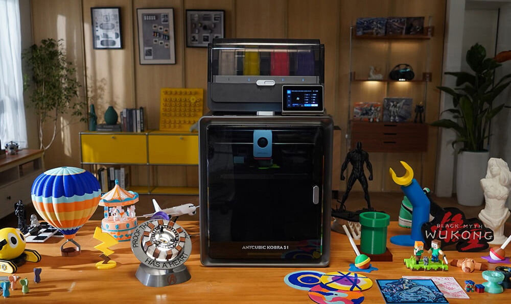 Na stole znajduje się drukarka 3D z napisem \'ANYCUBIC KOBRA S1\', otoczona różnorodnymi kolorowymi modelami, w tym balonem, karuzelą i figurkami. W tle widoczne są półki z książkami i dekoracjami oraz rośliny.