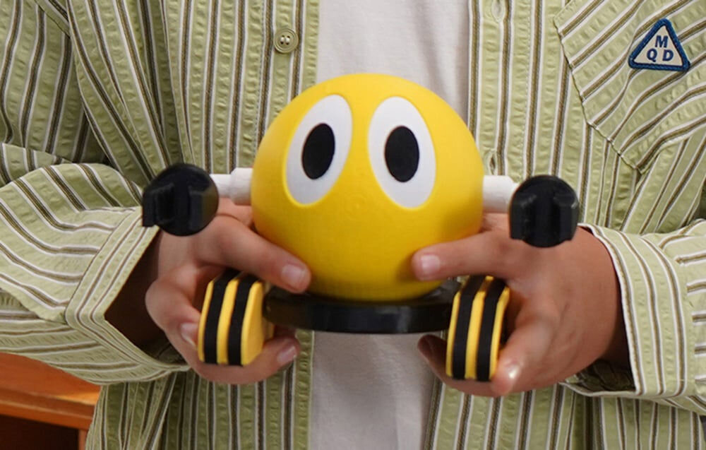 Osoba trzyma żółtą zabawkę z dużymi oczami i czarnymi elementami. Na koszuli widoczne jest logo z literami \'MQD\'.