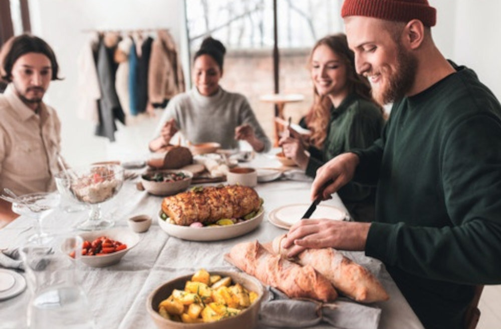 Grupa osób siedzi przy stole zastawionym różnorodnymi potrawami, w tym pieczywem i daniem mięsnym. W tle widać wieszak z ubraniami oraz duże okno.