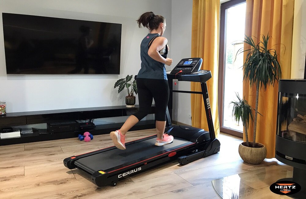 Osoba biegnie na bieżni elektrycznej z napisem \'CERRUS\' w jasnym pokoju z dużym oknem i roślinami. Na bieżni widoczne jest logo \'HERTZ\'.