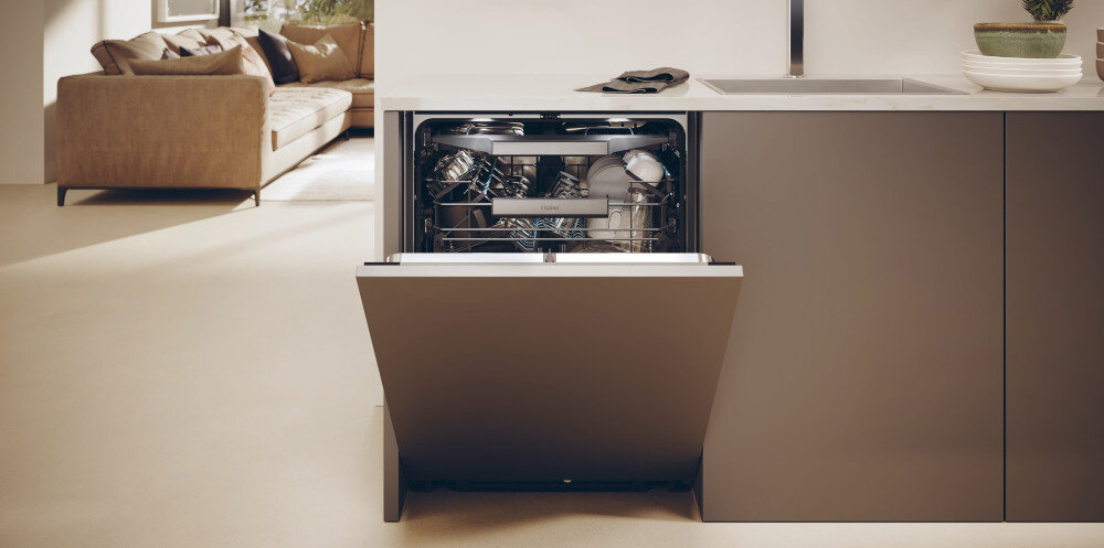 Otwarta zmywarka w zabudowie kuchennej z widocznymi naczyniami w środku. W tle znajduje się kanapa i roślina na blacie obok zlewu.