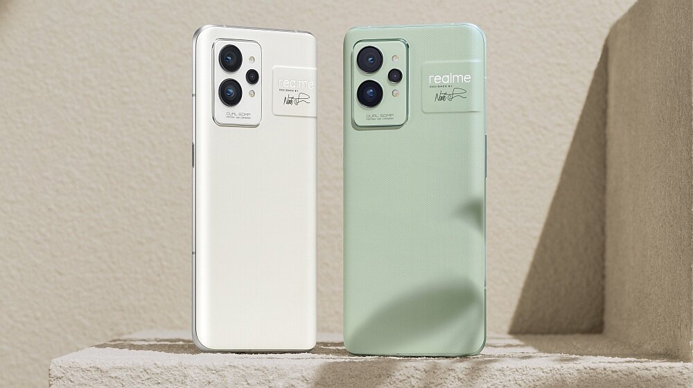Dwa smartfony w kolorach białym i zielonym stoją na jasnym tle. Na obu urządzeniach widoczne jest logo \'realme\' oraz podpis \'designed by Naoto Fukasawa\'.