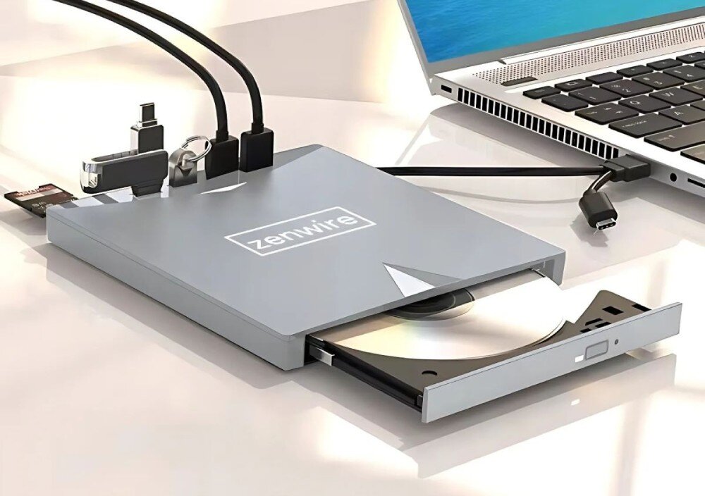 Na obrazie widoczny jest zewnętrzny napęd optyczny z logo \'ZenWire\', podłączony do laptopa. Napęd ma otwartą tackę z płytą CD, a obok znajdują się różne kable i adaptery.