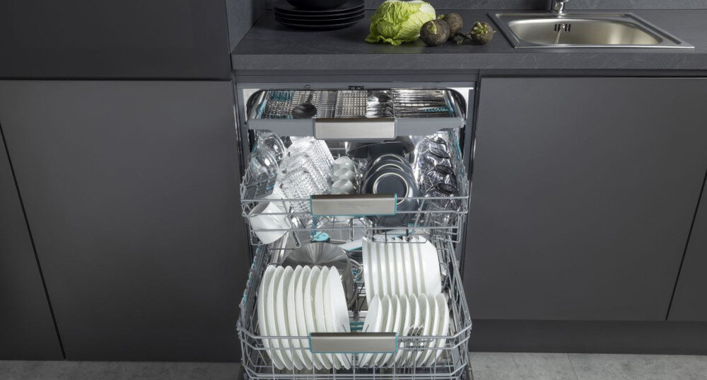 Zmywarka z otwartymi drzwiami, wypełniona naczyniami, sztućcami i szklankami na dwóch poziomach koszy. W tle widoczny jest blat kuchenny z umywalką i warzywami.