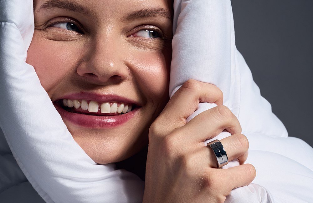 Osoba owinięta w biały materiał uśmiecha się, trzymając rękę przy twarzy. Na palcu widoczny jest czarny pierścień.