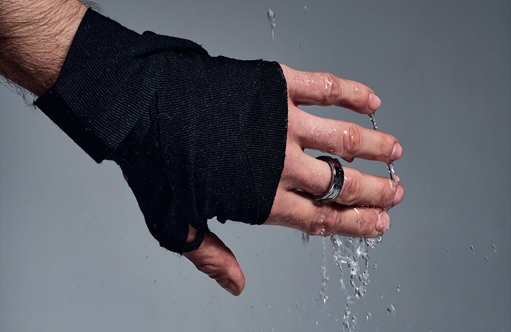 Ręka owinięta czarnym materiałem, na palcu znajduje się czarny pierścień, a woda spływa po dłoni.
