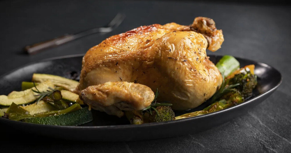 Na czarnym talerzu znajduje się pieczony kurczak z warzywami, takimi jak cukinia i brokuły. W tle widoczny jest rozmazany widelec na ciemnym blacie.