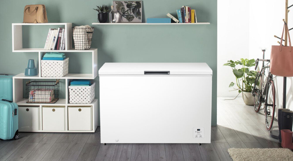Biała zamrażarka skrzyniowa stoi w pomieszczeniu z drewnianą podłogą, obok regału z książkami i ozdobami. W tle widać rower oraz rośliny doniczkowe.