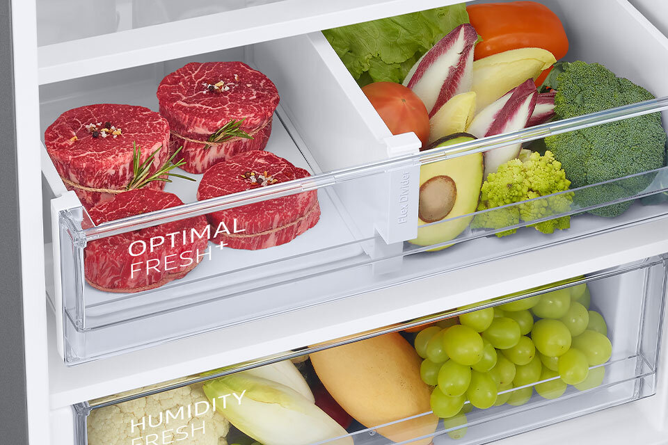 Wnętrze lodówki z dwoma szufladami, w jednej z nich znajdują się kawałki mięsa z napisem \'OPTIMAL FRESH+\', a w drugiej różne warzywa i owoce. Widoczne są m.in. brokuły, awokado, winogrona i pomidory.