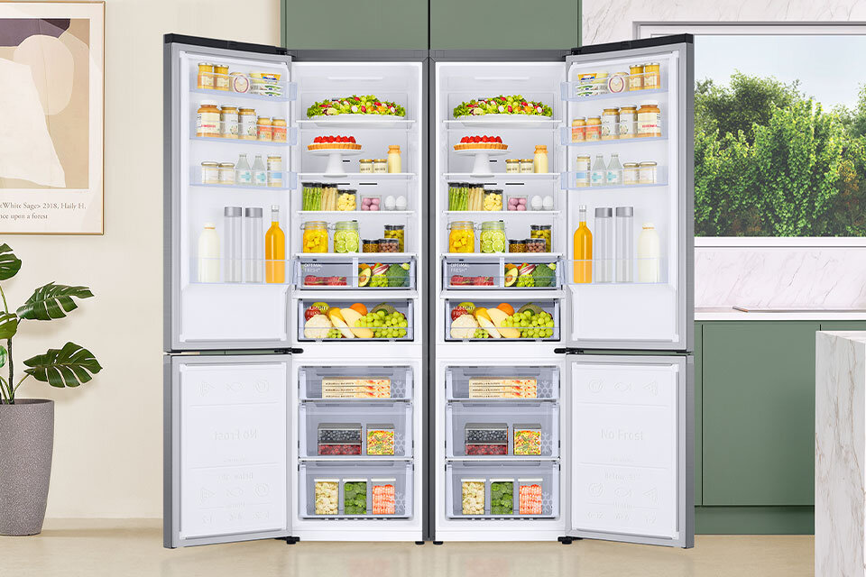 Dwie otwarte lodówki wypełnione różnorodnymi produktami spożywczymi, takimi jak owoce, warzywa, napoje i słoiki. W tle widoczne są zielone szafki kuchenne, roślina doniczkowa oraz okno z widokiem na zieleń.