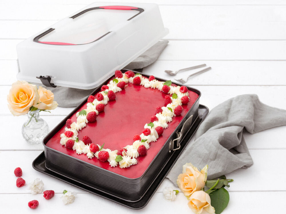 Na obrazie znajduje się prostokątna forma do pieczenia z ciastem ozdobionym bitą śmietaną i malinami. Obok formy leży szara serwetka, sztućce oraz wazon z żółtymi różami.