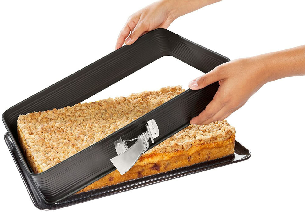 Na obrazie widoczna jest prostokątna forma do pieczenia z metalowym zapięciem, trzymana przez dwie ręce. W formie znajduje się ciasto z kruszonką na wierzchu.