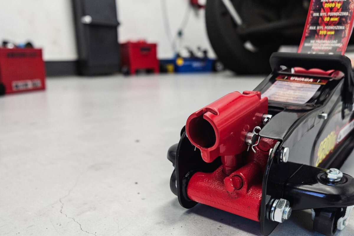 Na obrazie widoczny jest podnośnik hydrauliczny w kolorze czarno-czerwonym, ustawiony na jasnej podłodze w warsztacie. W tle znajdują się rozmyte narzędzia i sprzęty warsztatowe.