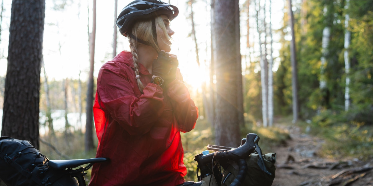 Osoba w czerwonej kurtce i kasku rowerowym stoi obok roweru w lesie, z widocznym zegarkiem na nadgarstku. W tle widać drzewa i słońce przebijające się przez gałęzie.