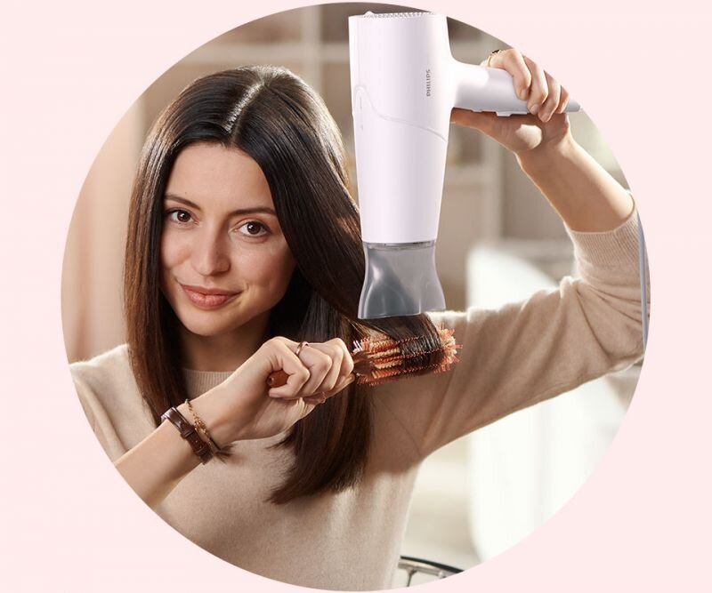 Osoba używa białej suszarki do włosów marki Philips, stylizując włosy na okrągłej szczotce. W tle widoczne jest rozmyte wnętrze pomieszczenia.