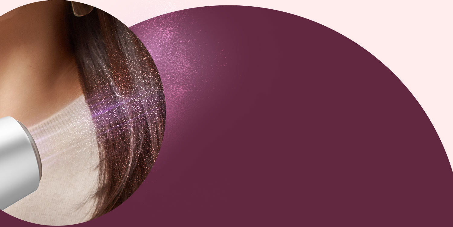 Obraz przedstawia fragment suszarki do włosów skierowanej na ciemne, lśniące włosy. Tło jest w odcieniach fioletu i różu.