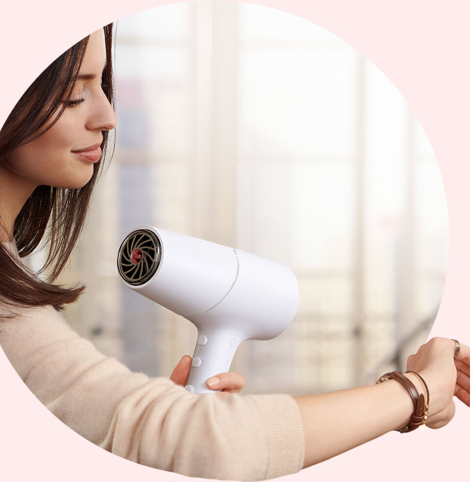 Osoba trzyma białą suszarkę do włosów, skierowaną w stronę włosów. W tle widoczne jest rozmyte wnętrze pomieszczenia.