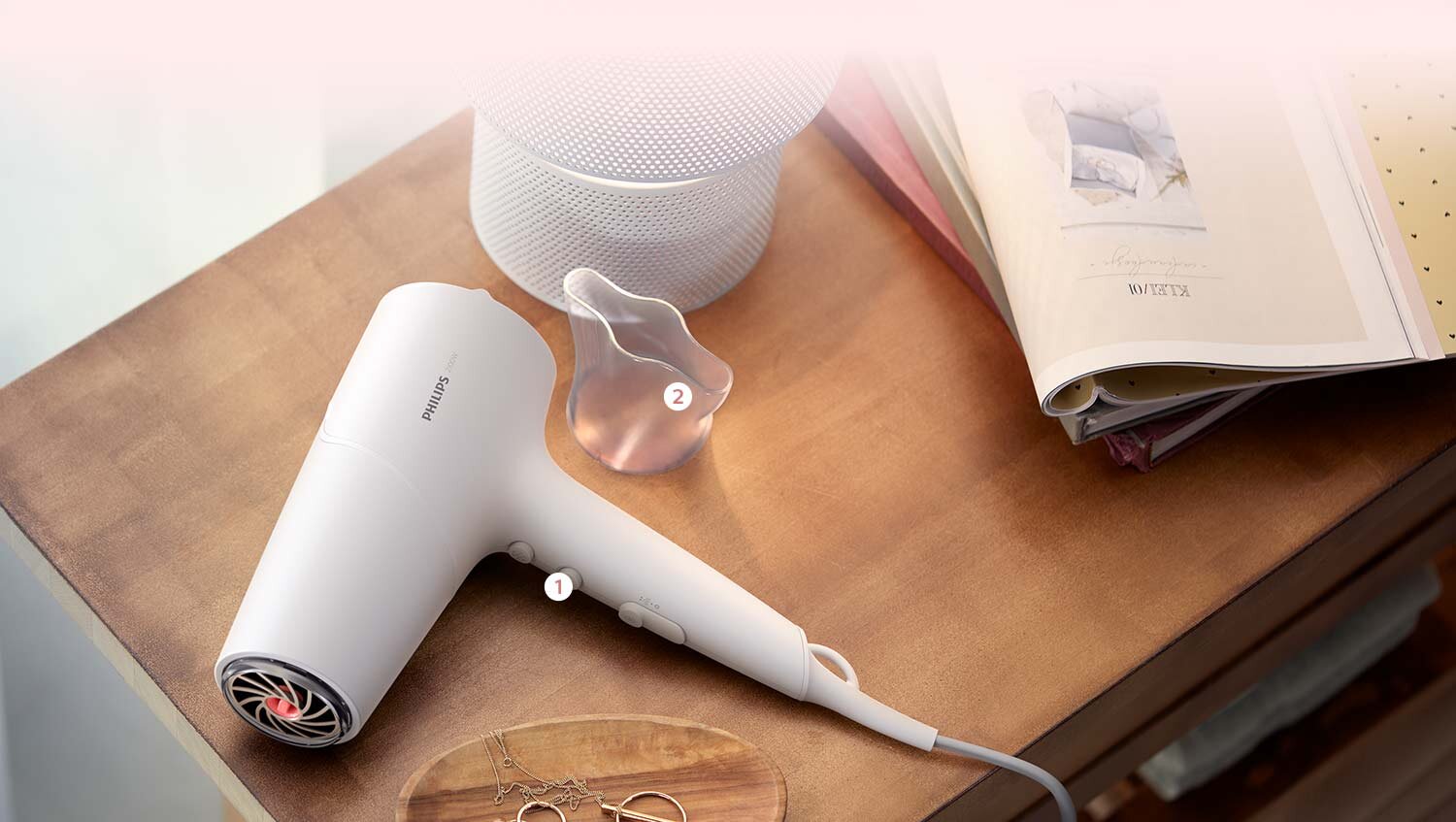 Na drewnianym stole leży biała suszarka z widocznym logo \'PHILIPS\', obok której znajduje się przezroczysta nasadka. W tle otwarta książka i biały głośnik.