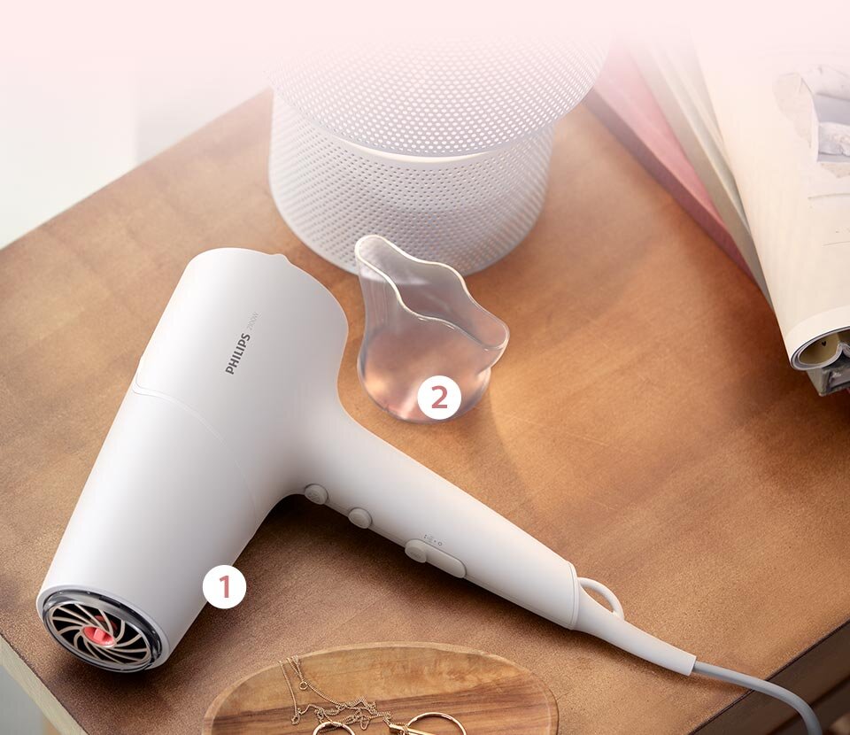 Na drewnianym stole leży biała suszarka do włosów z widocznym logo \'PHILIPS\'. Obok znajduje się przezroczysta nasadka oraz otwarta książka.