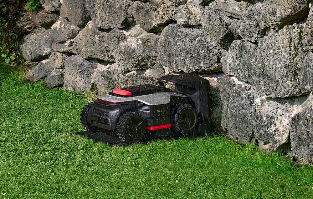 Robot koszący o ciemnej obudowie z czerwonymi akcentami znajduje się na trawie obok kamiennego muru. Urządzenie ma widoczne duże koła i kompaktową konstrukcję.
