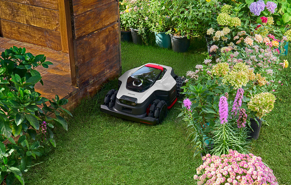 Robot koszący porusza się po trawniku otoczonym kwiatami i drewnianą konstrukcją. W tle widoczne są różnorodne rośliny w doniczkach.