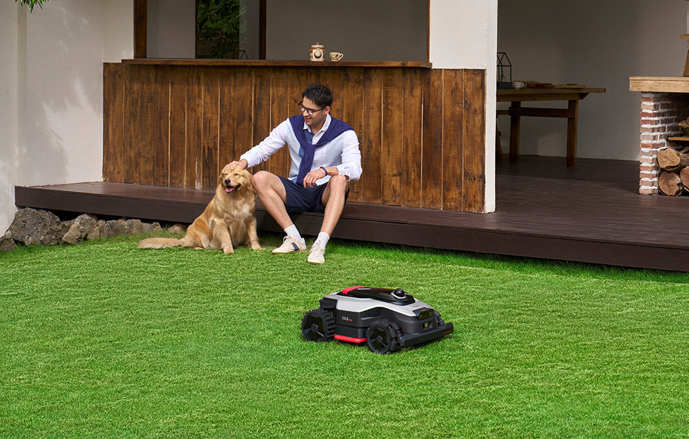 Na trawniku znajduje się robot koszący, a w tle siedzi mężczyzna z psem na drewnianym tarasie.