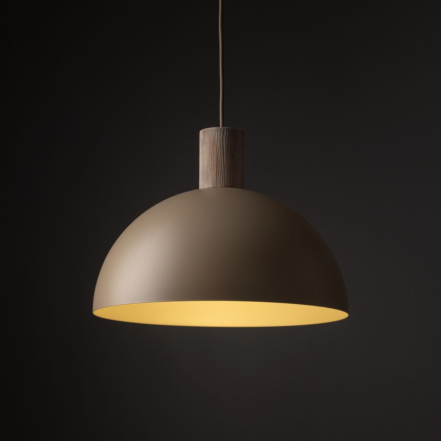 Na obrazie widoczna jest beżowa, metalowa lampa wisząca o półkulistym kształcie z drewnianym elementem na górze. Tło jest ciemne, co podkreśla jasne wnętrze klosza.