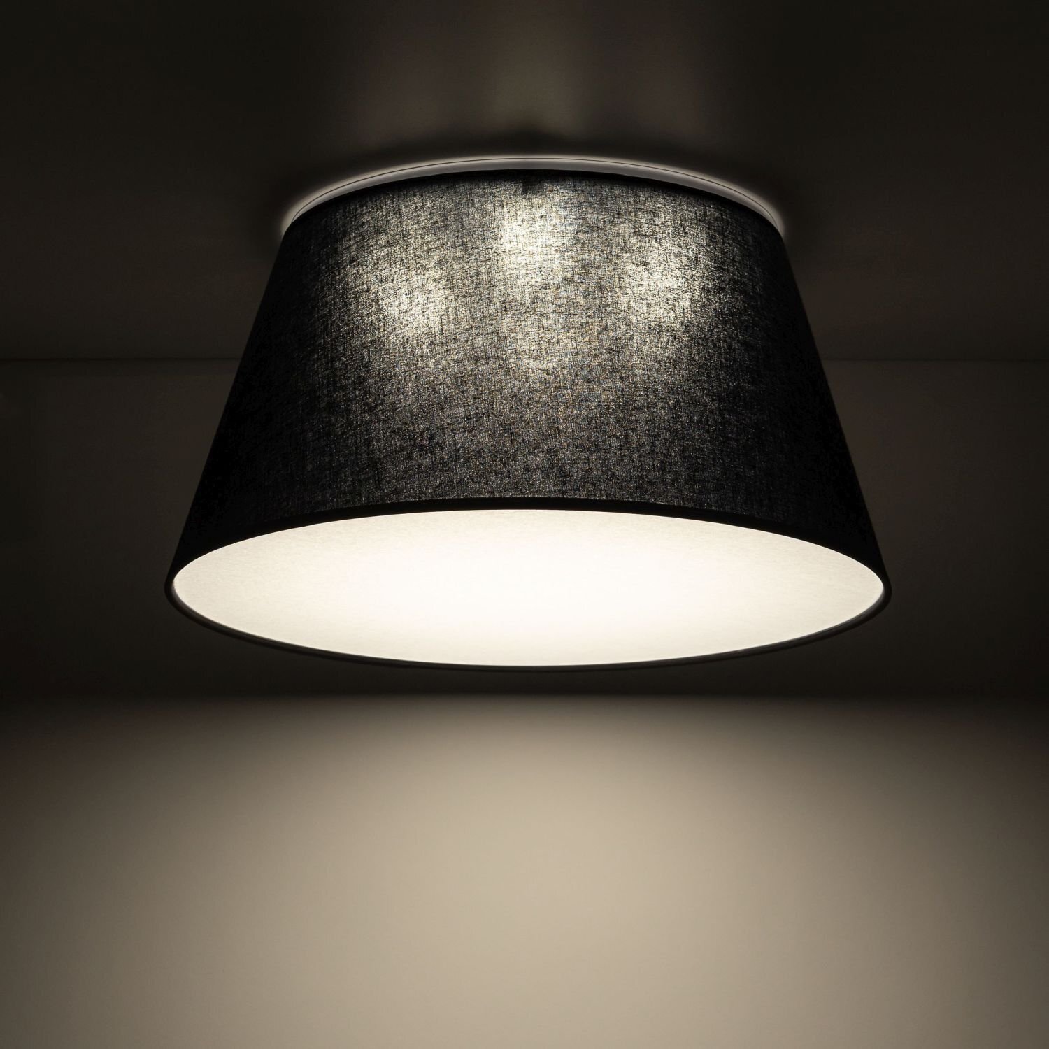 Czarna lampa sufitowa o stożkowym kształcie z białym wnętrzem, emitująca ciepłe światło. Lampa jest zamontowana na suficie w ciemnym pomieszczeniu.