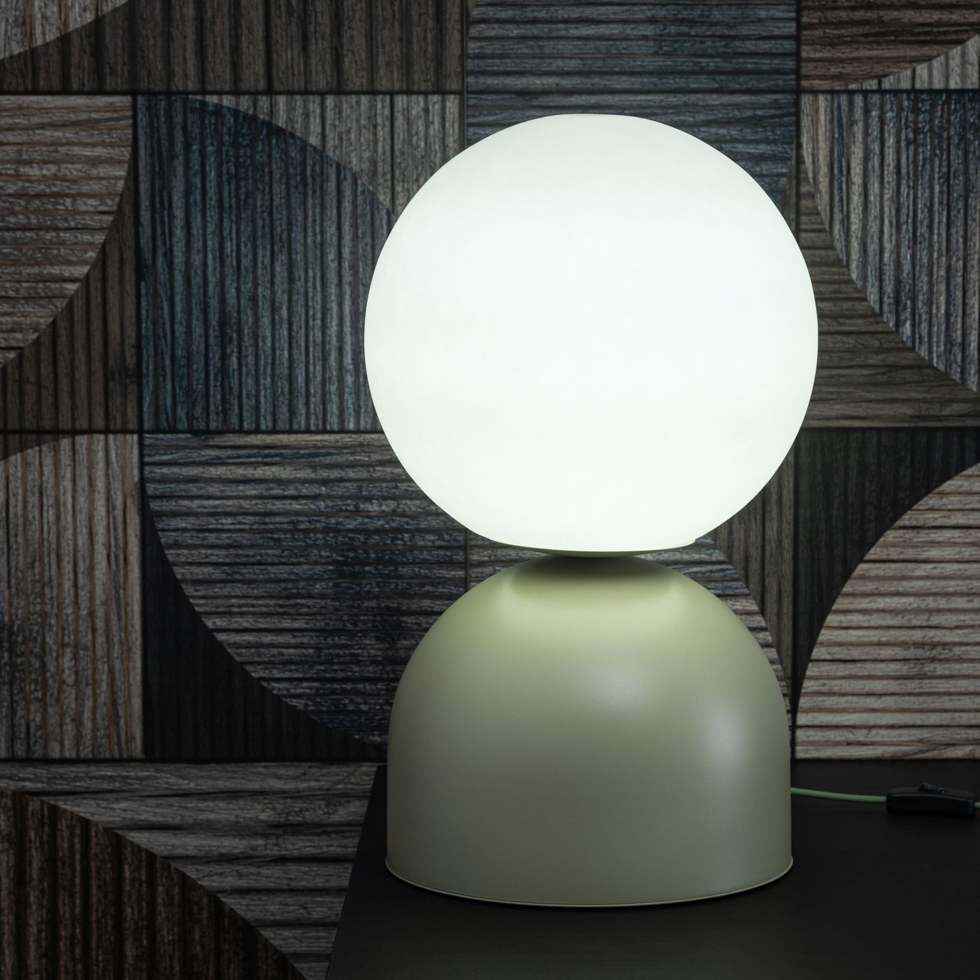 Na zdjęciu znajduje się nowoczesna lampka nocna z pistacjową, lakierowaną podstawą i kulistym kloszem. W tle widoczna jest ściana z geometrycznym wzorem w odcieniach szarości.