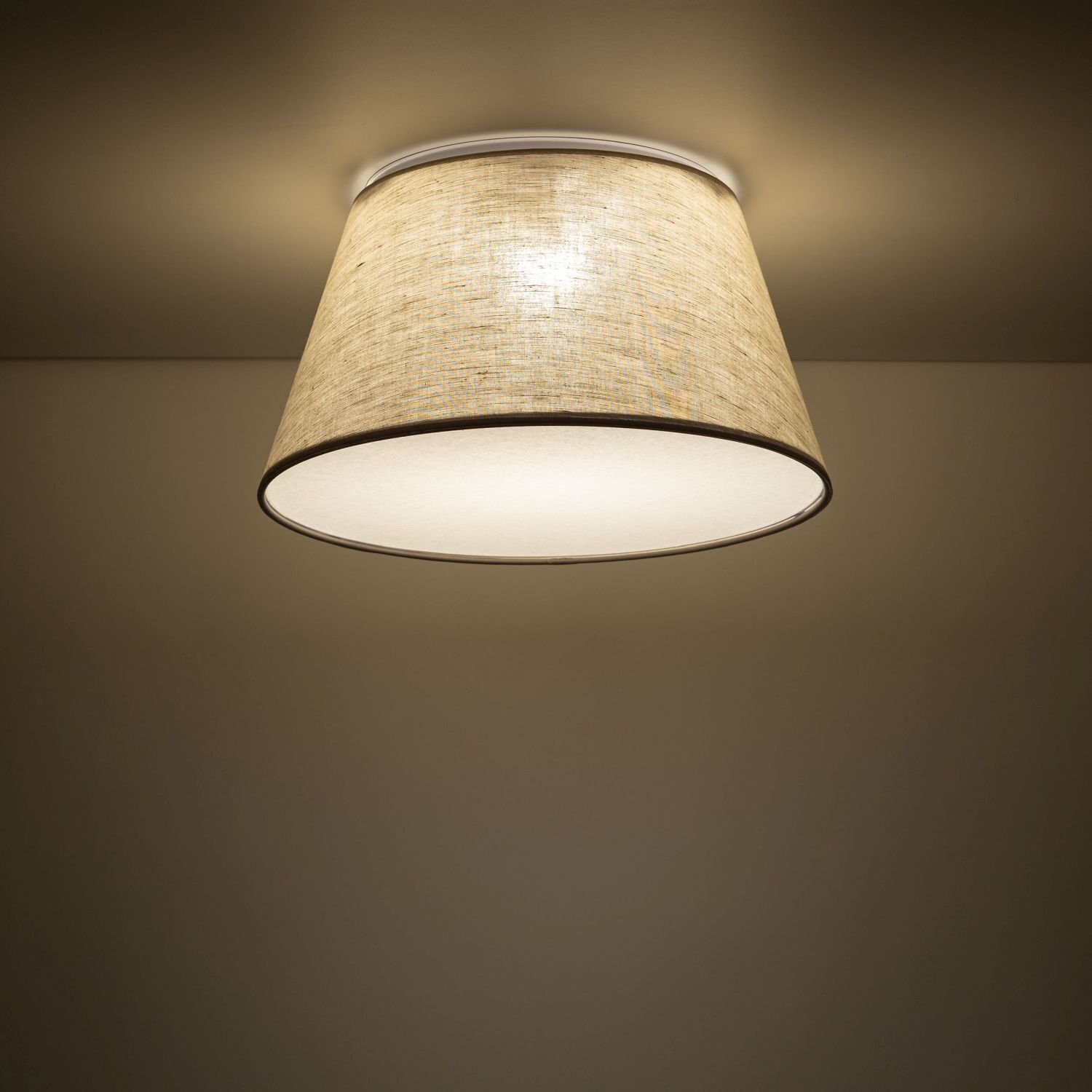 Na obrazie widoczna jest lampa sufitowa z beżowym, tekstylnym kloszem o stożkowym kształcie. Światło emituje ciepłą poświatę na sufit i ściany.