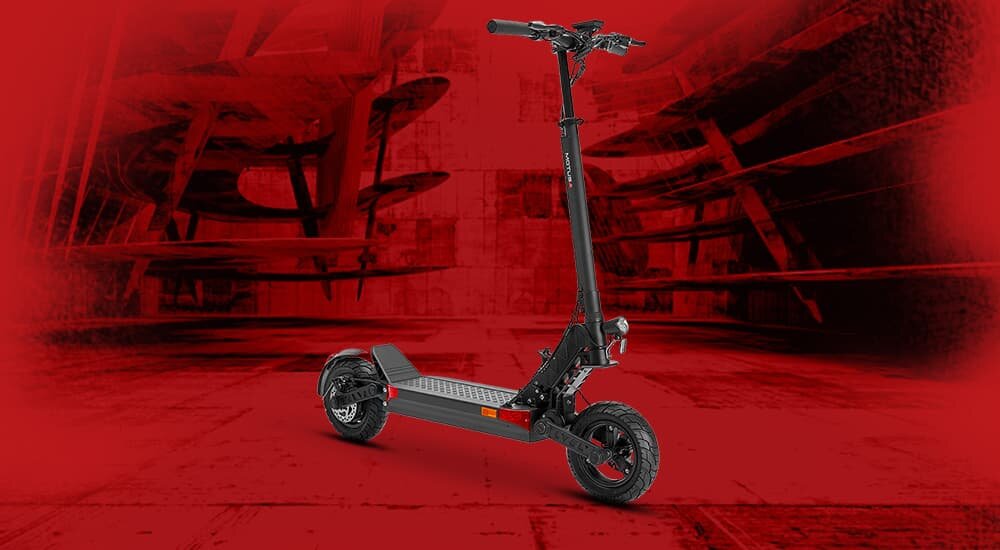 Czarna hulajnoga elektryczna z dużymi kołami stoi na tle czerwonego, industrialnego wnętrza. Hulajnoga ma solidną konstrukcję i widoczne amortyzatory.