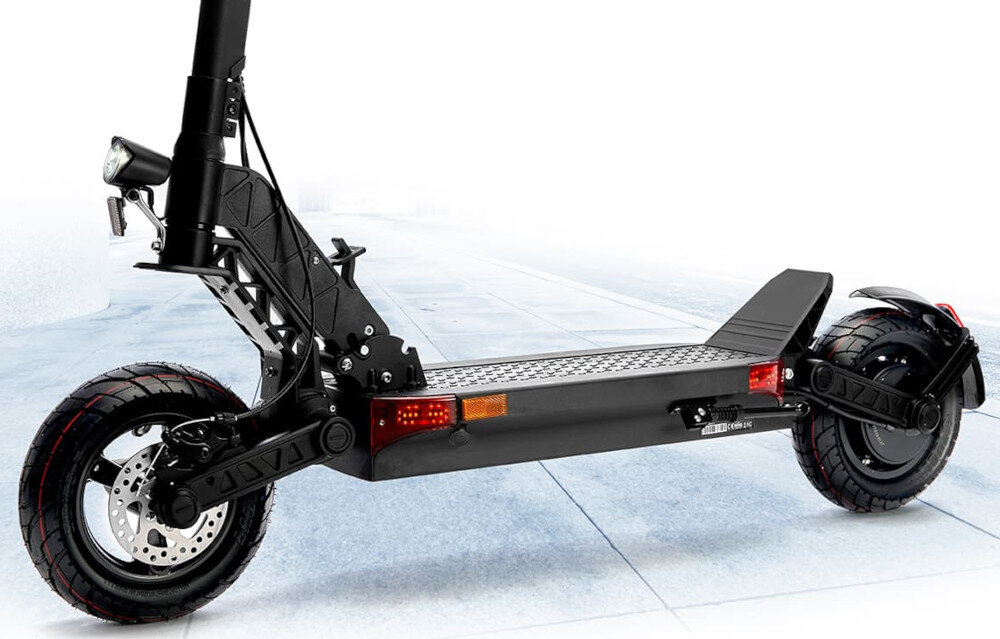 Czarna hulajnoga elektryczna z dużymi oponami i amortyzacją, widoczna z boku na tle chodnika. Na podeście znajduje się wzór kratki, a z przodu zamontowane jest światło.