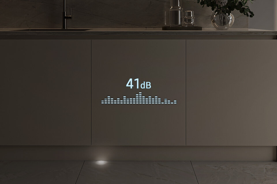 Na froncie szafki kuchennej wyświetlany jest napis \'41 dB\' oraz graficzny wskaźnik poziomu dźwięku. W tle widoczny jest zlew i dekoracja z rośliną.