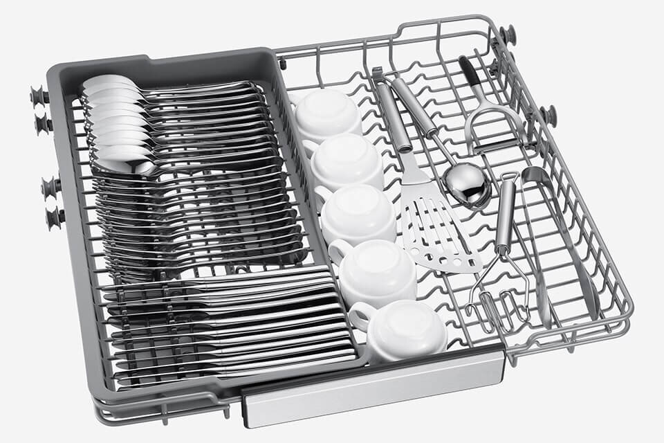 Wnętrze zmywarki z ułożonymi sztućcami i filiżankami na specjalnej tacy. Widoczne są łyżki, widelce, noże oraz kilka białych filiżanek.