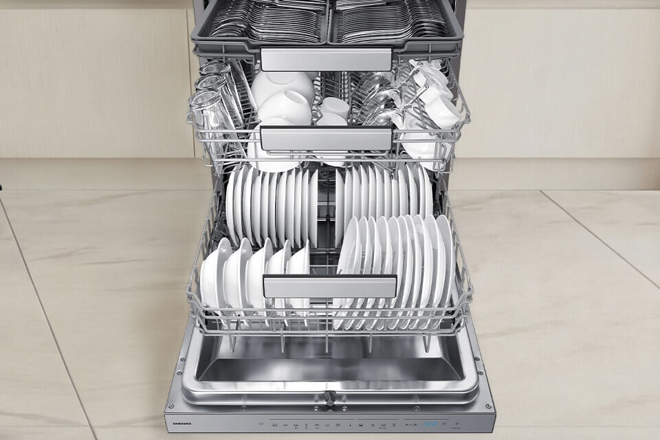 Zmywarka z trzema poziomami koszy, wypełniona naczyniami, takimi jak talerze, kubki i sztućce. Na panelu widoczne jest logo \'SAMSUNG\'.