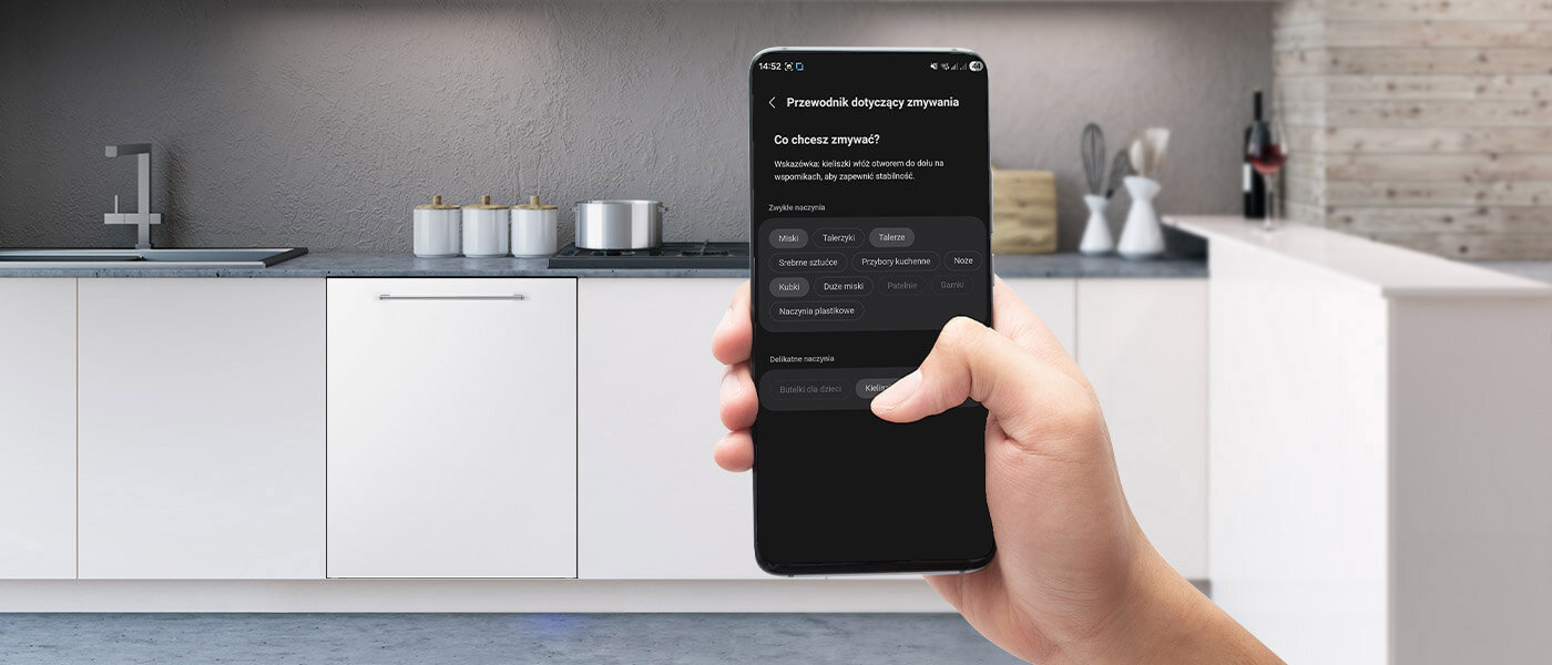 Osoba trzyma smartfon z aplikacją dotyczącą zmywania, na tle nowoczesnej kuchni z białymi szafkami i zlewem. Na ekranie telefonu widoczny jest tekst: \'Przewodnik dotyczący zmywania\' oraz opcje wyboru naczyń do zmywania.