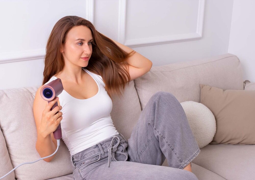 Osoba siedzi na kanapie, trzymając różową suszarkę do włosów. Na wyświetlaczu suszarki widoczna jest liczba \'25\'.