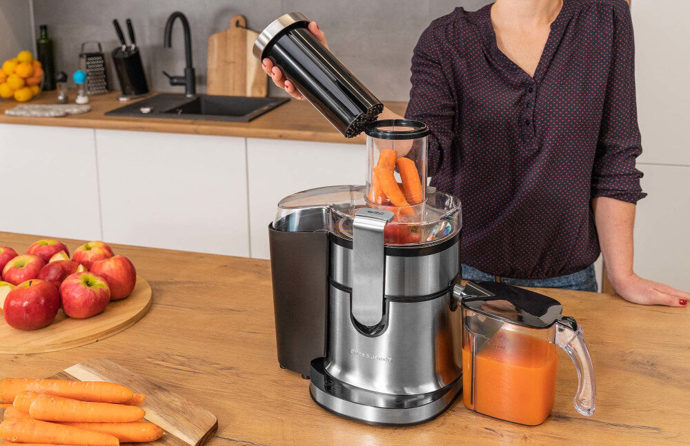 Na blacie kuchennym stoi sokowirówka z logo \'GÖTZE & JENSEN\', do której wkładane są marchewki. Obok znajduje się dzbanek z sokiem, a w tle widać jabłka i zlew.