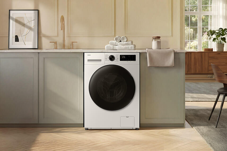 Pralko-suszarka Samsung WD90HG5U34BEEO w ofercie Media Expert Pralko-suszarka w zabudowie kuchennej z białym frontem i czarnym bębnem. Na blacie znajdują się złożone ręczniki i dekoracje.