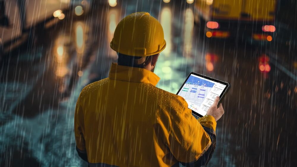 Osoba w żółtym ubraniu ochronnym trzyma tablet, na którym widoczny jest interfejs aplikacji. Pada deszcz, a w tle widać rozmyte światła pojazdów.