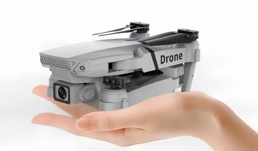 Na zdjęciu widać złożony dron w kolorze szarym z napisem \'Drone\', trzymany na dłoni. Dron ma widoczną kamerę z przodu.