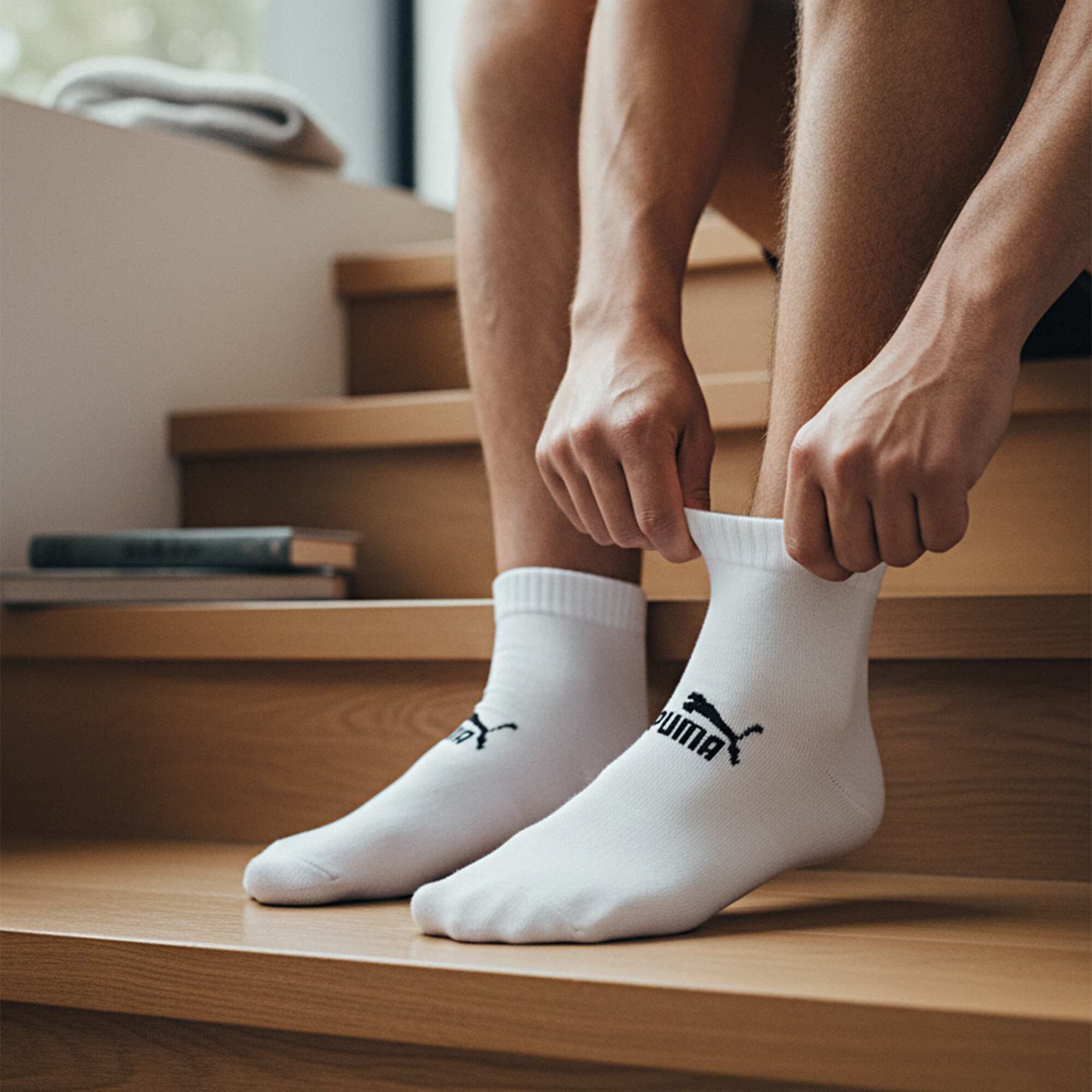 Osoba siedzi na schodach i zakłada białe skarpety z logo Puma. W tle widoczne są książki i ręcznik.