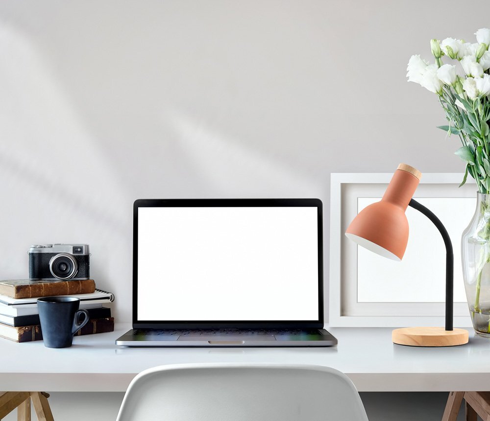 Na biurku znajduje się laptop z białym ekranem, obok którego stoi lampa biurkowa o pomarańczowym kloszu i czarnym ramieniu. W tle widoczny jest wazon z białymi kwiatami, aparat fotograficzny oraz stos książek.