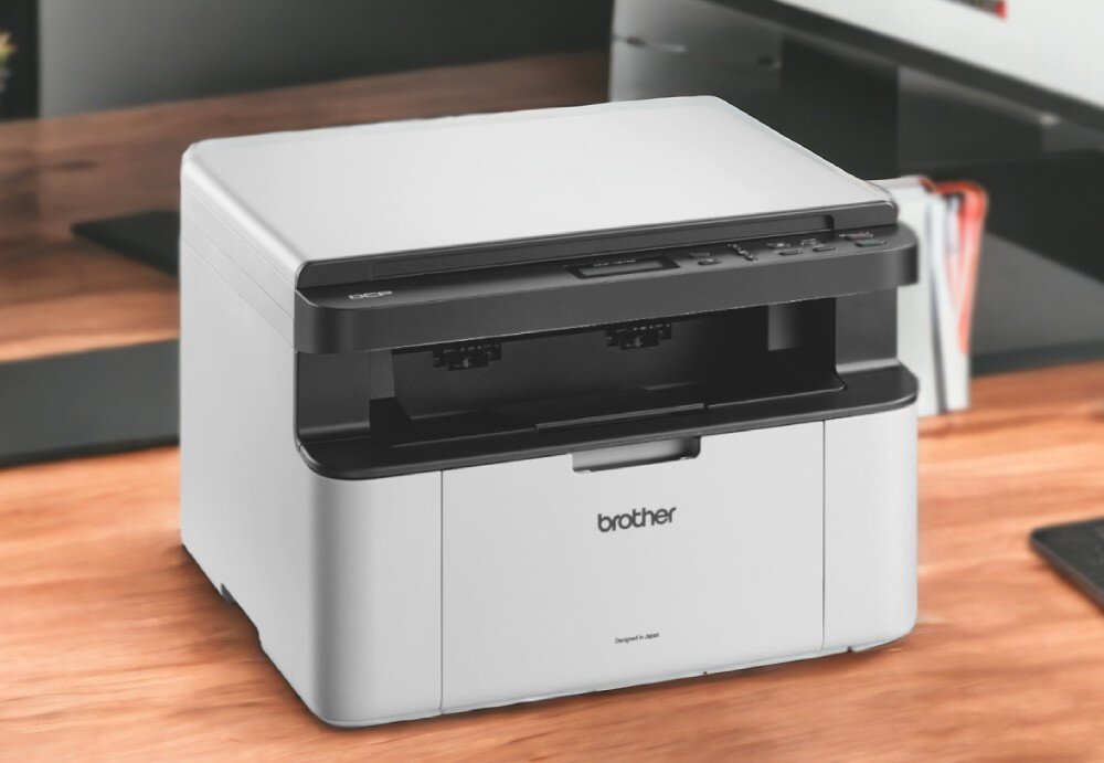 Na drewnianym biurku stoi urządzenie wielofunkcyjne w kolorze białym z czarnymi elementami, z widocznym logo \'brother\' na przedniej części. W tle widać fragmenty biurka i inne elementy biurowe.