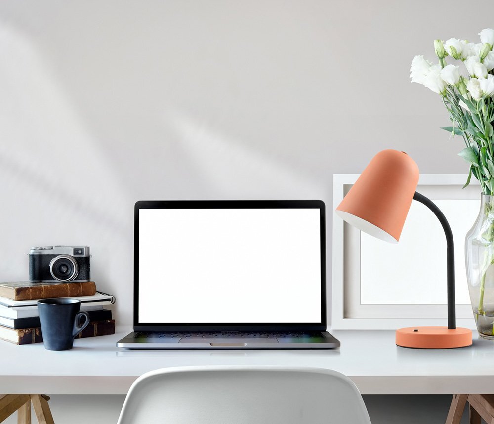Na biurku znajduje się laptop z białym ekranem, obok którego stoi pomarańczowa lampa biurkowa z czarnym ramieniem. W tle widać aparat fotograficzny, stos książek, kubek oraz wazon z białymi kwiatami.