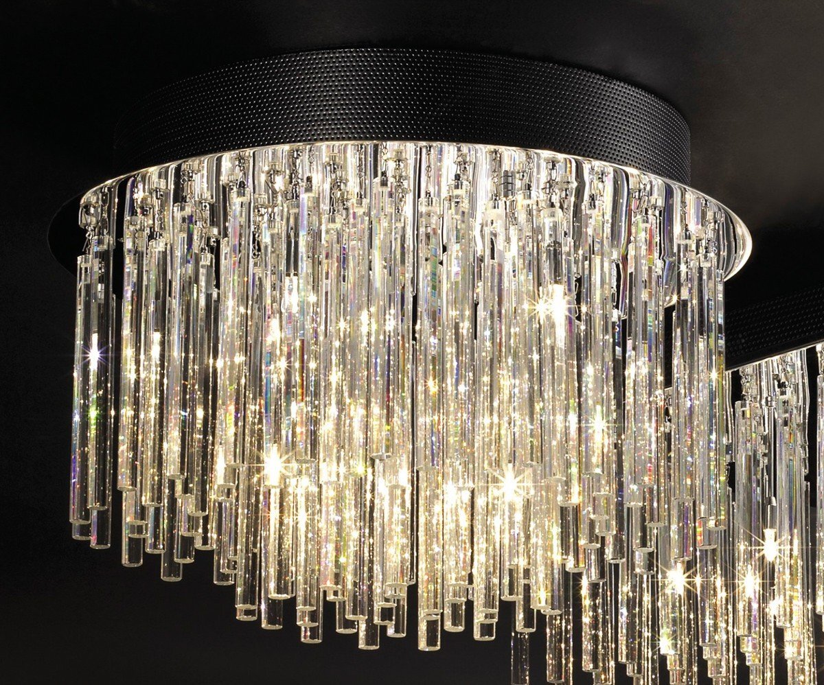 Lampa sufitowa złożona z licznych, pionowych, szklanych elementów, które odbijają światło, tworząc efektowne refleksy. Górna część lampy jest czarna i ma teksturowaną powierzchnię.