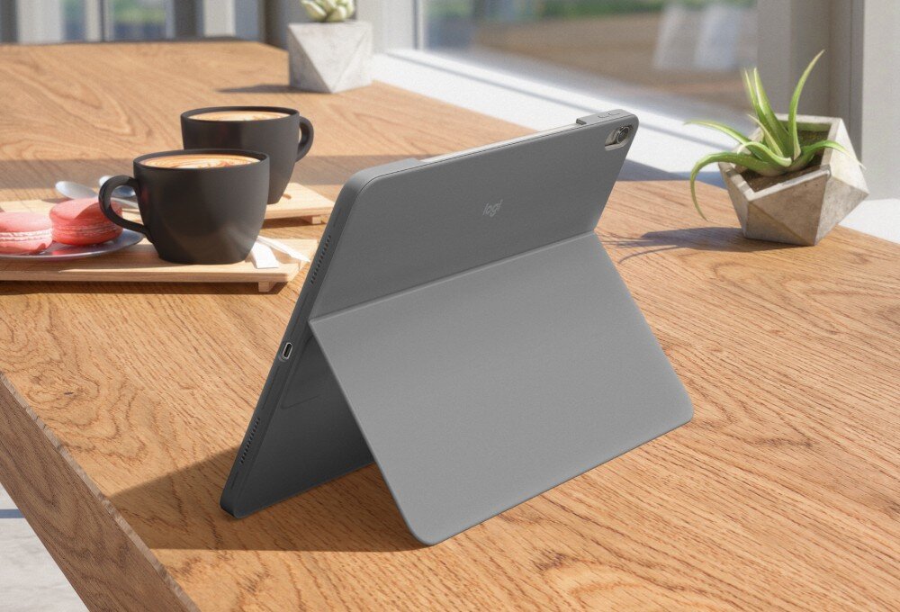 Na drewnianym stole stoi szare etui na tablet z klawiaturą, ustawione w pozycji stojącej. Obok znajdują się dwie filiżanki z napojem oraz talerz z makaronikami, a w tle widać roślinę w doniczce.